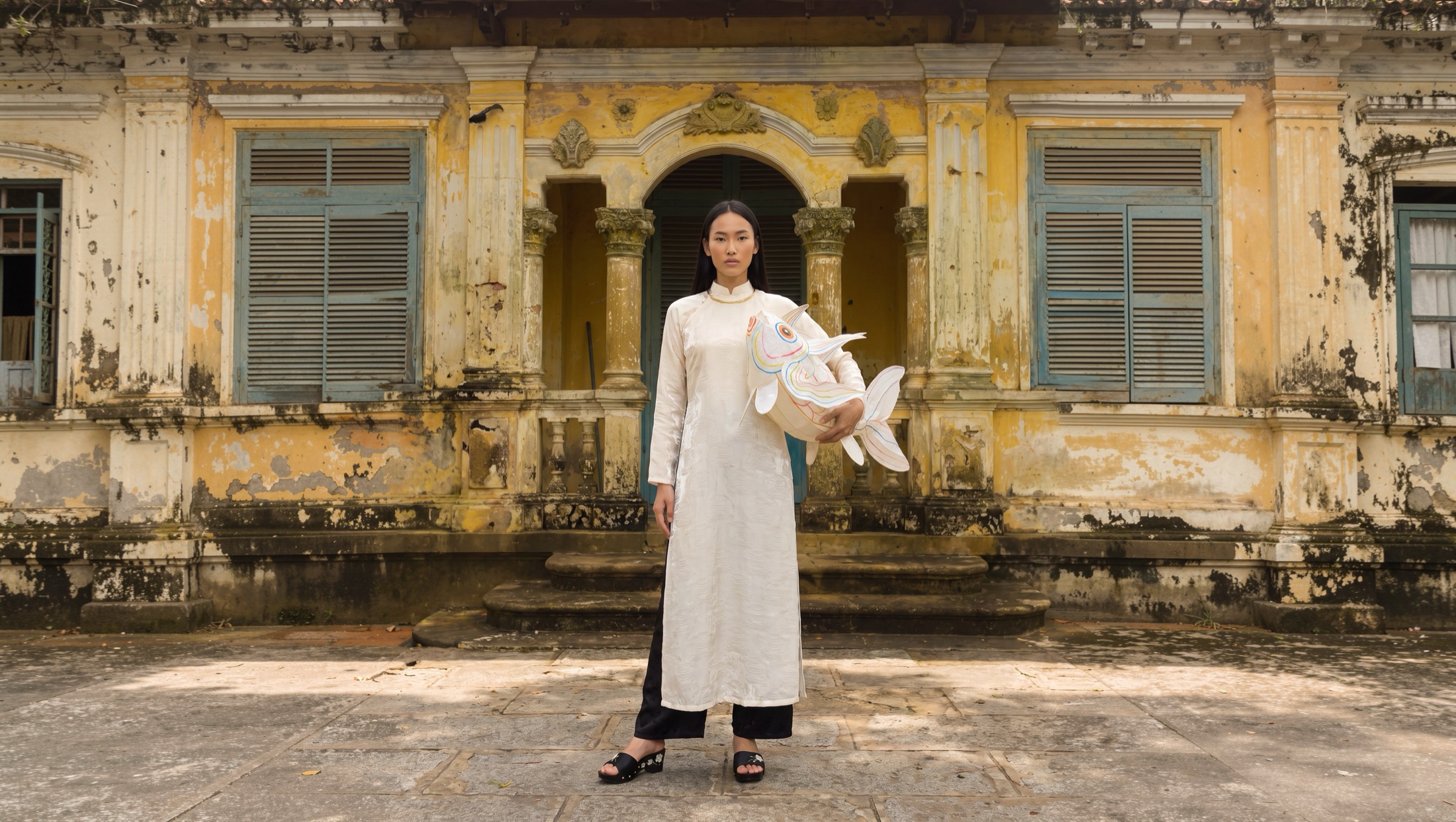 A model in a white áo dài holding a paper fish lantern in front of Nhà Cổ Huỳnh Công Ngữ, a century-old house in Long An